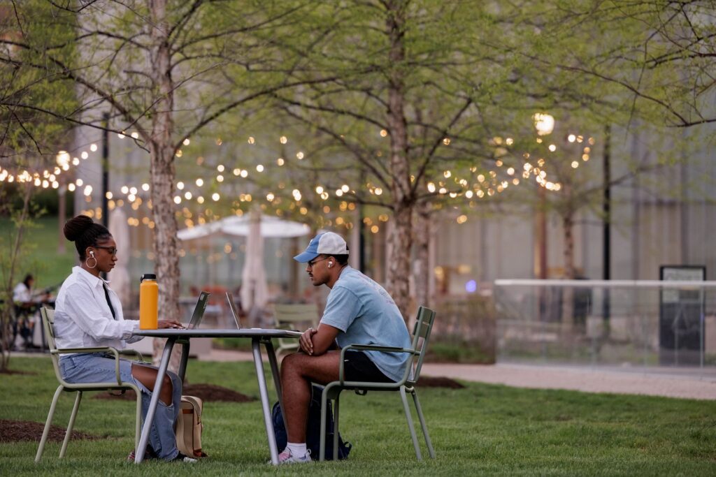 Two students sit at an outdoor table looking at their laptops with trees and string lights behind them