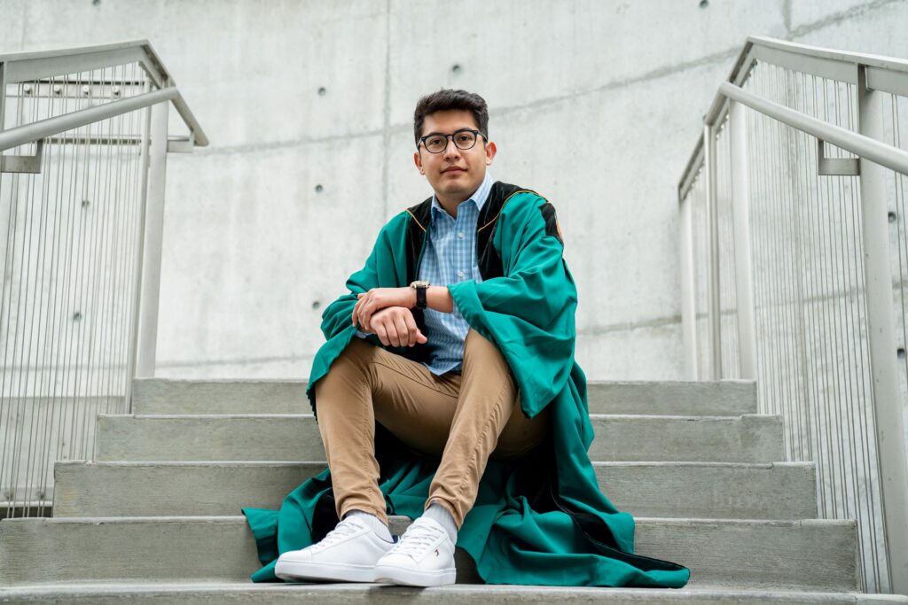 Student with glasses dressed in green WashU graduation gown sits on stairs