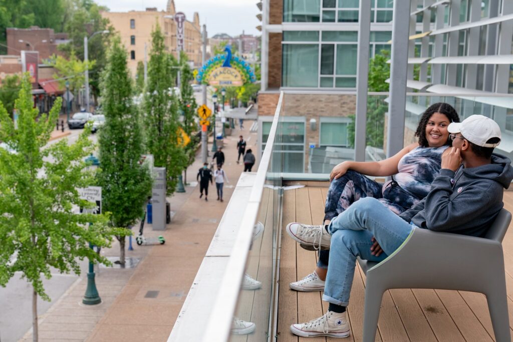 Two students sit on the outdoor balcony of the Lofts building overlooking people walking on the street below