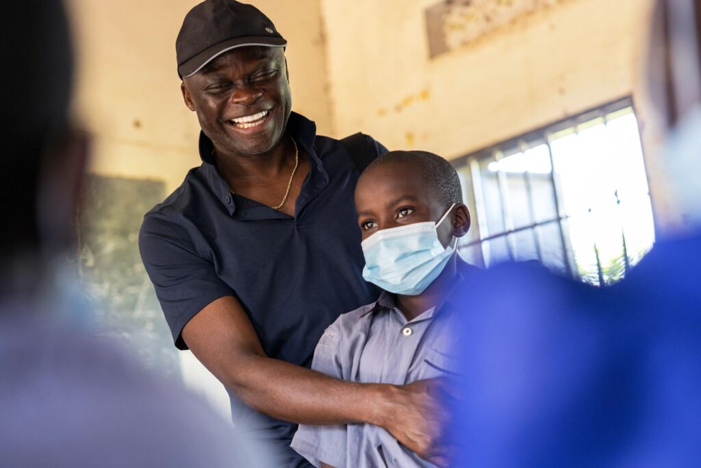 Professor Fred M. Ssewamala and a child with a mask on stand in a classroom in Uganda