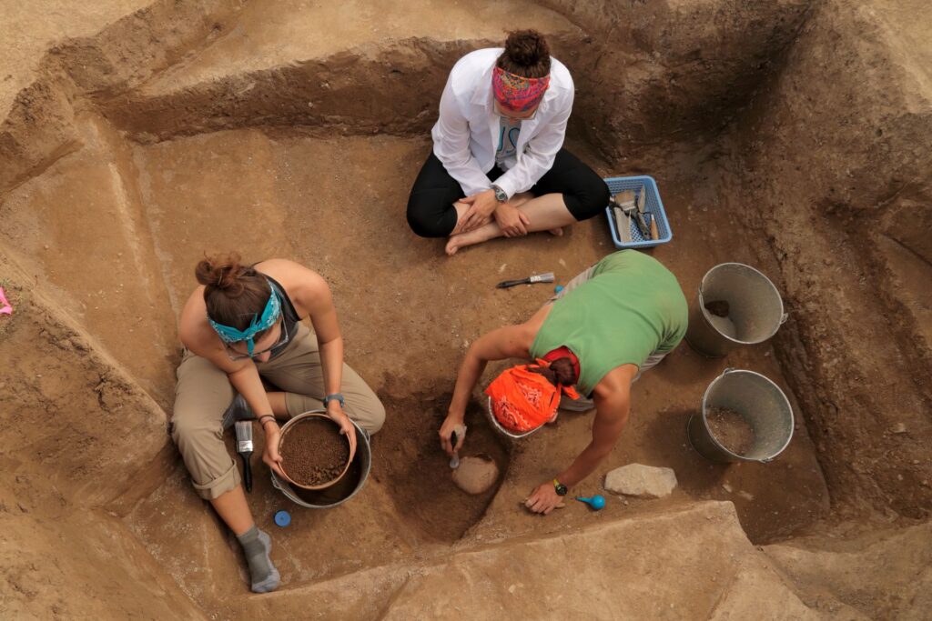 Three archeologists sit on the ground and use excavation tools to brush dirt away from a potential artifact