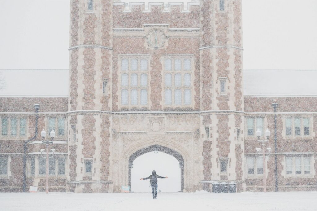 A student stands in front of Brookings Hall with snow in the air and on the ground around them