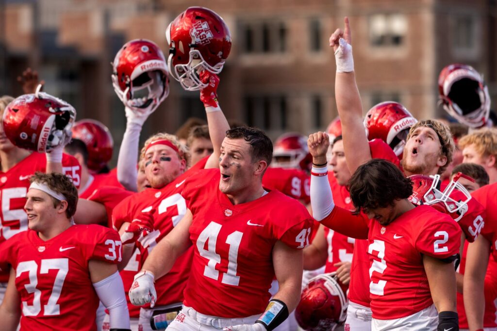 WashU Bears football players in red and white uniforms cheer and hold their helmets up