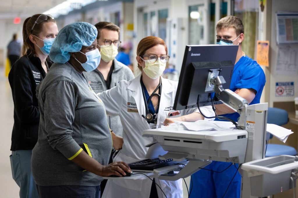 Doctors, nurses and other staff with medical clothes and masks on gather around a computer screen within a hospital