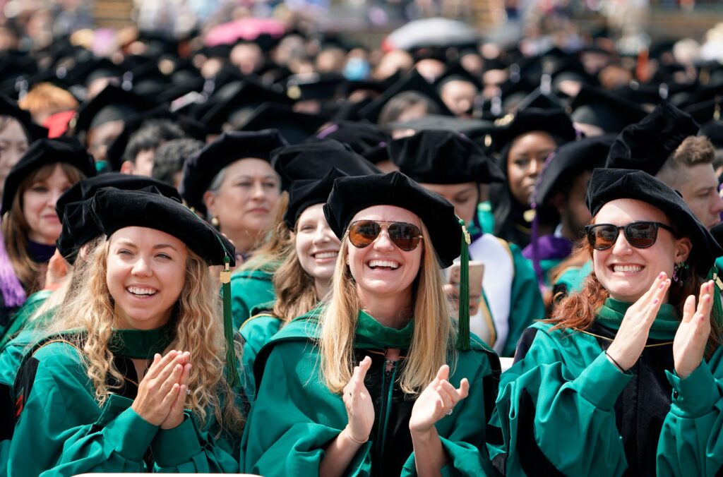 Crowd of graduates in their green gowns and black caps clap and cheer while watching graduation ceremony