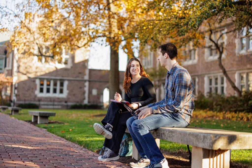 Undergraduate students Annabel Rose (left) and Sam Sweat study together on a bench with fall foliage in the background